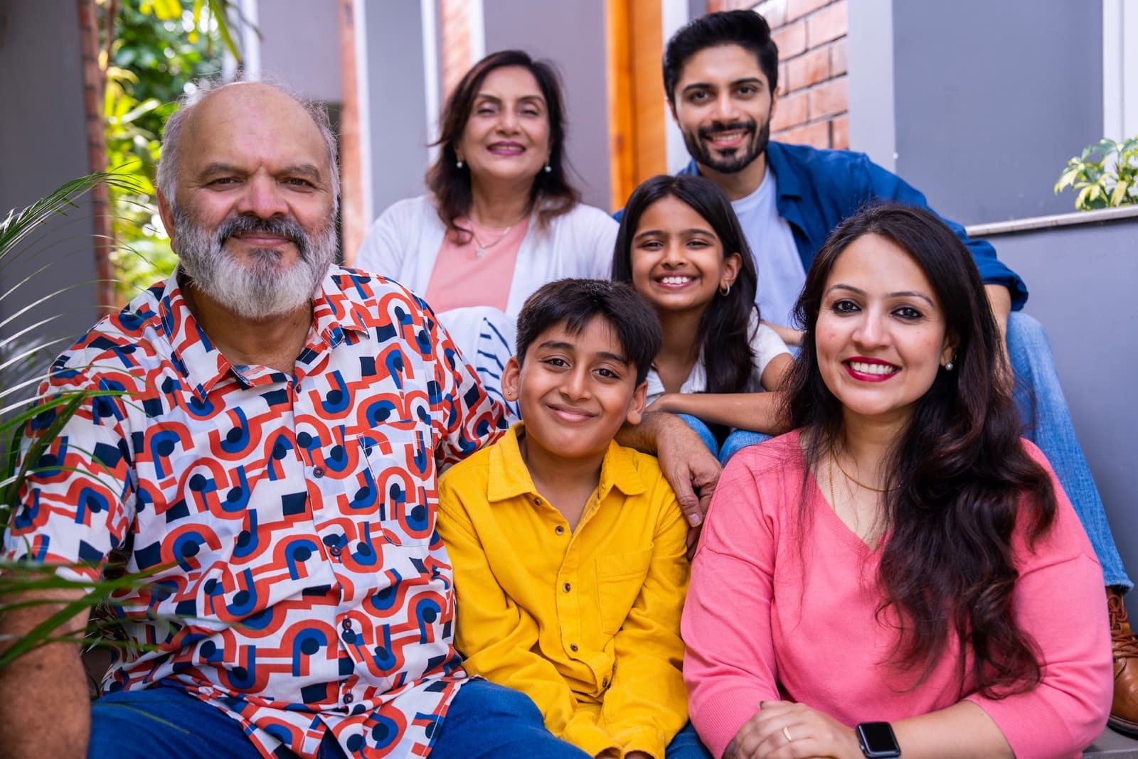 Three generations of an Indian family — grandparents, parents and two children — sitting together and smiling on their home steps