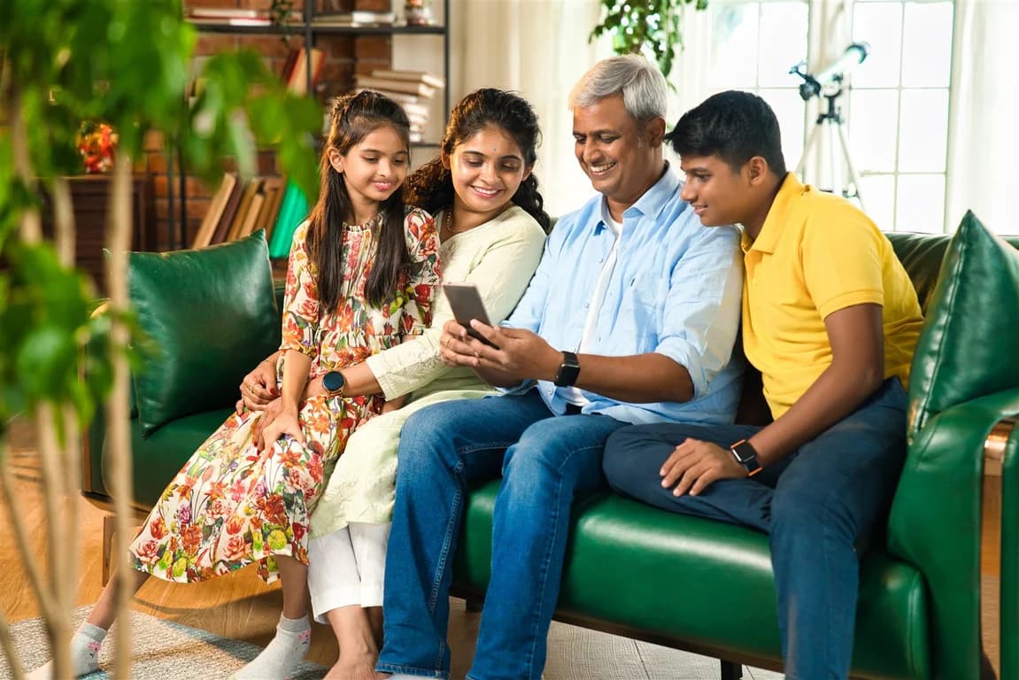 An Indian family of four sitting together on a sofa, smiling while looking at a health app on a smartphone